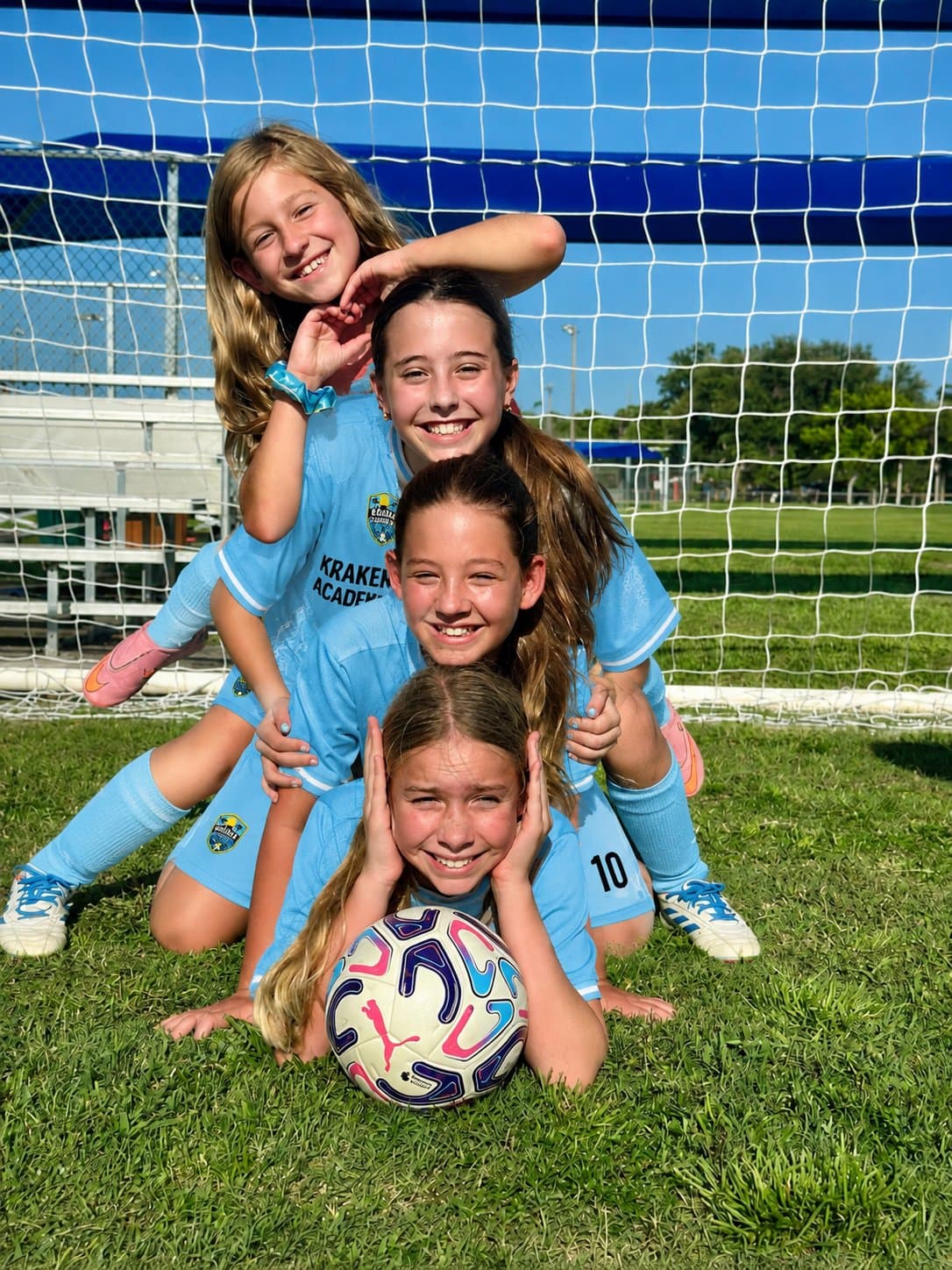 Four Kraken Academy players stacked in a pyramid on the grass, the player in front resting her chin on a ball