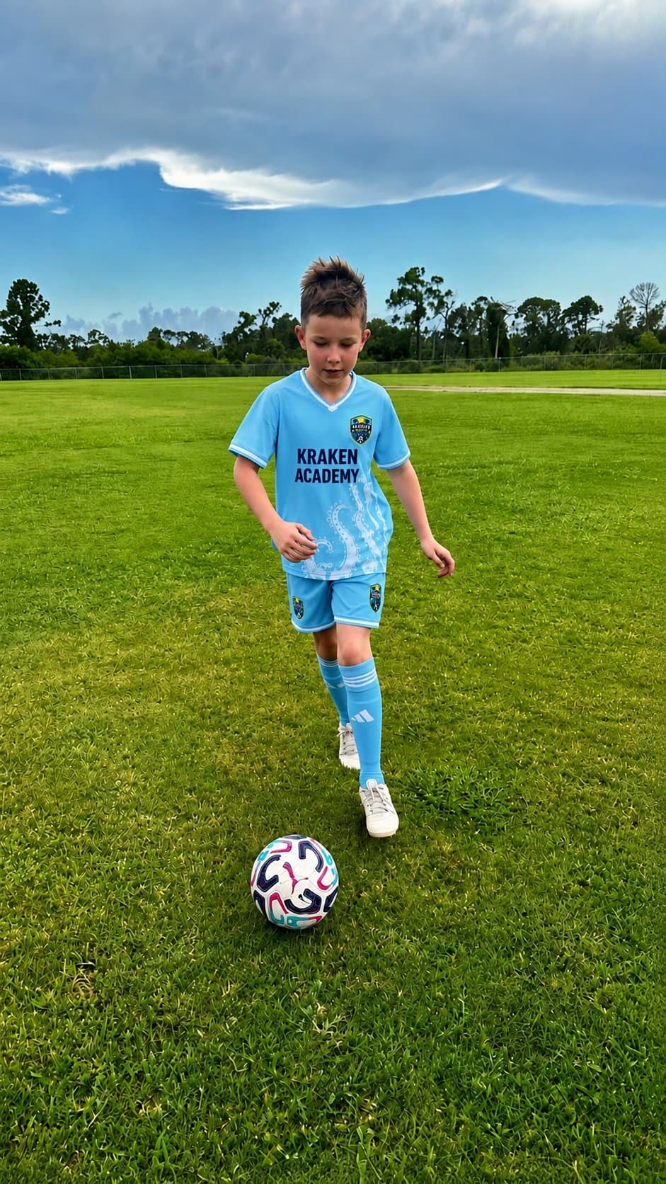 A Kraken Academy player dribbling a ball across open grass under a heavy sky