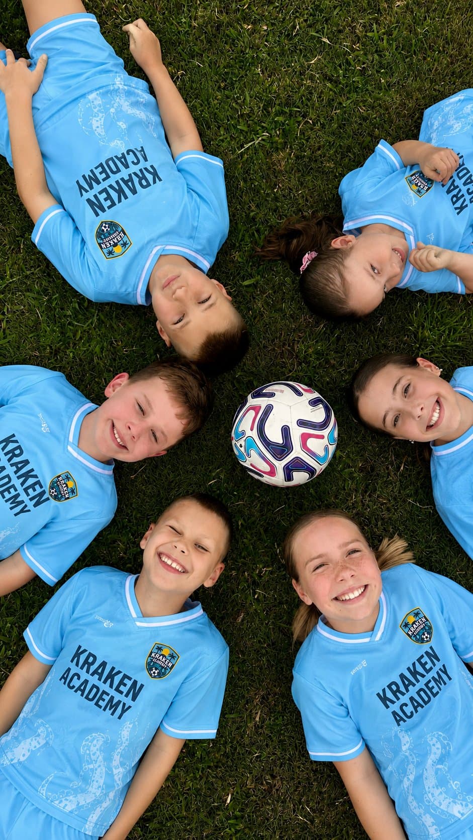 Seven Kraken Academy players lying on the grass in a circle with their heads together and a ball at the centre, photographed from above