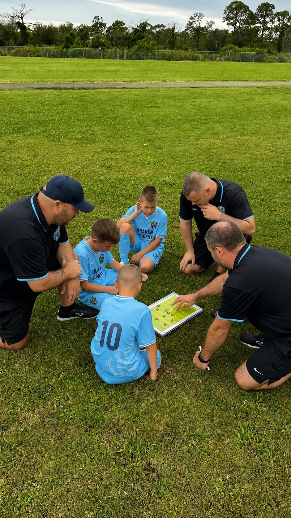 Three Kraken Academy coaches crouched with young players around a tactics board on the pitch
